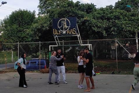 Gruppenspielende Menschen auf einem Basketballplatz, im Hintergrund eine &bdquo;ULTRAS&ldquo;-Bande und B&auml;ume unter blauem Himmel.