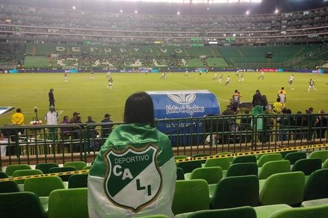 Fan mit Deportivo Cali-Jacke sitzt in einem Stadion und beobachtet ein Fu&szlig;ballspiel auf einem gr&uuml;nen Rasenplatz.