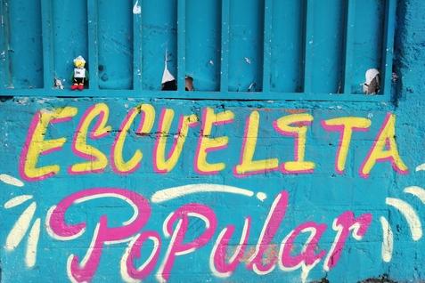 Bunte Wand mit der Beschriftung &bdquo;ESCUELITA POPULAR&ldquo; und kleinen Fig&uuml;rchen zwischen den Gitterst&auml;ben.