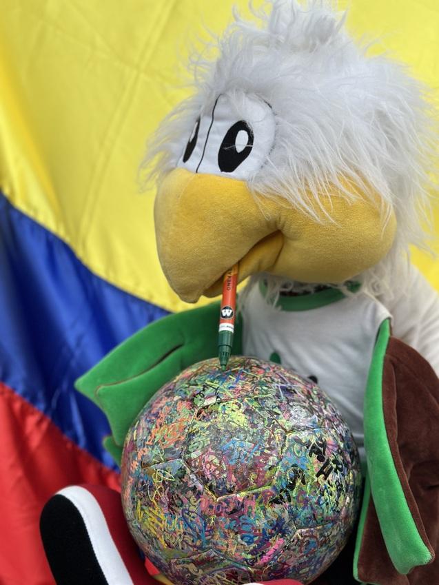 Kuscheltier in Form eines Vogels h&auml;lt einen Stift und sitzt neben einem bunten Ball vor einer tri-farbigen Fahne.