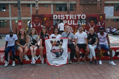 Gruppe von acht Personen posiert mit einem Banner vor einer farbenfrohen Wand, beschriftet mit &bdquo;DISTRITO POPULAR&ldquo;.