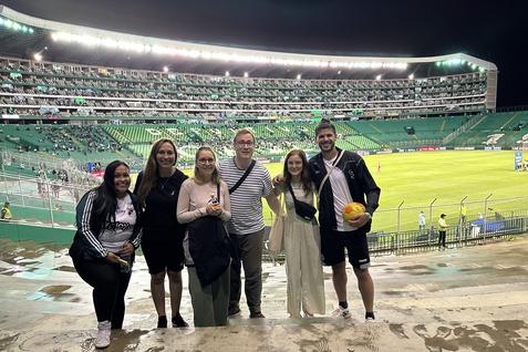 Gruppe von sechs Personen posiert vor einem Fu&szlig;ballstadion mit gr&uuml;ner Trib&uuml;ne und Spielfeld im Hintergrund.