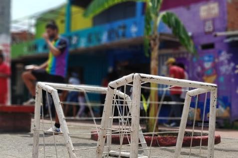 Zwei kleine, wei&szlig;e Fu&szlig;balltore stehen auf einem gepflasterten Boden vor bunten H&auml;usern und einer Palme.