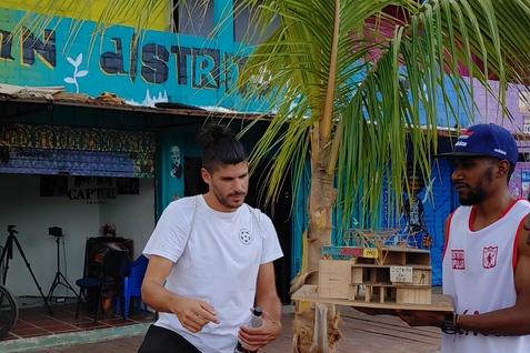 Mann in wei&szlig;em T-Shirt steht neben einer Palme und h&auml;lt eine Flasche, w&auml;hrend ein anderer Mann ein Holzobjekt &uuml;berreicht.
