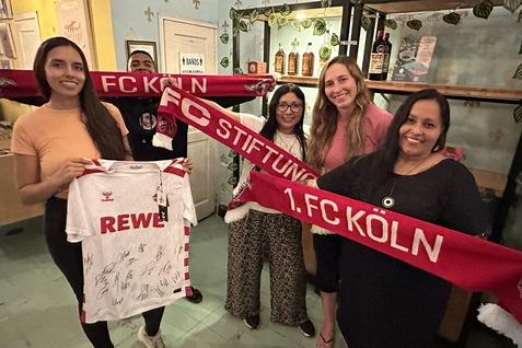 F&uuml;nf Personen halten FC K&ouml;ln-Schals und ein signiertes Trikot in einem geschm&uuml;ckten Raum.