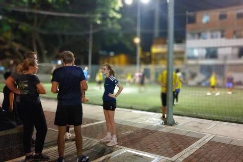 Gruppe junger Menschen steht am Rand eines Fu&szlig;ballplatzes, w&auml;hrend im Hintergrund ein Spiel bei Nacht stattfindet.