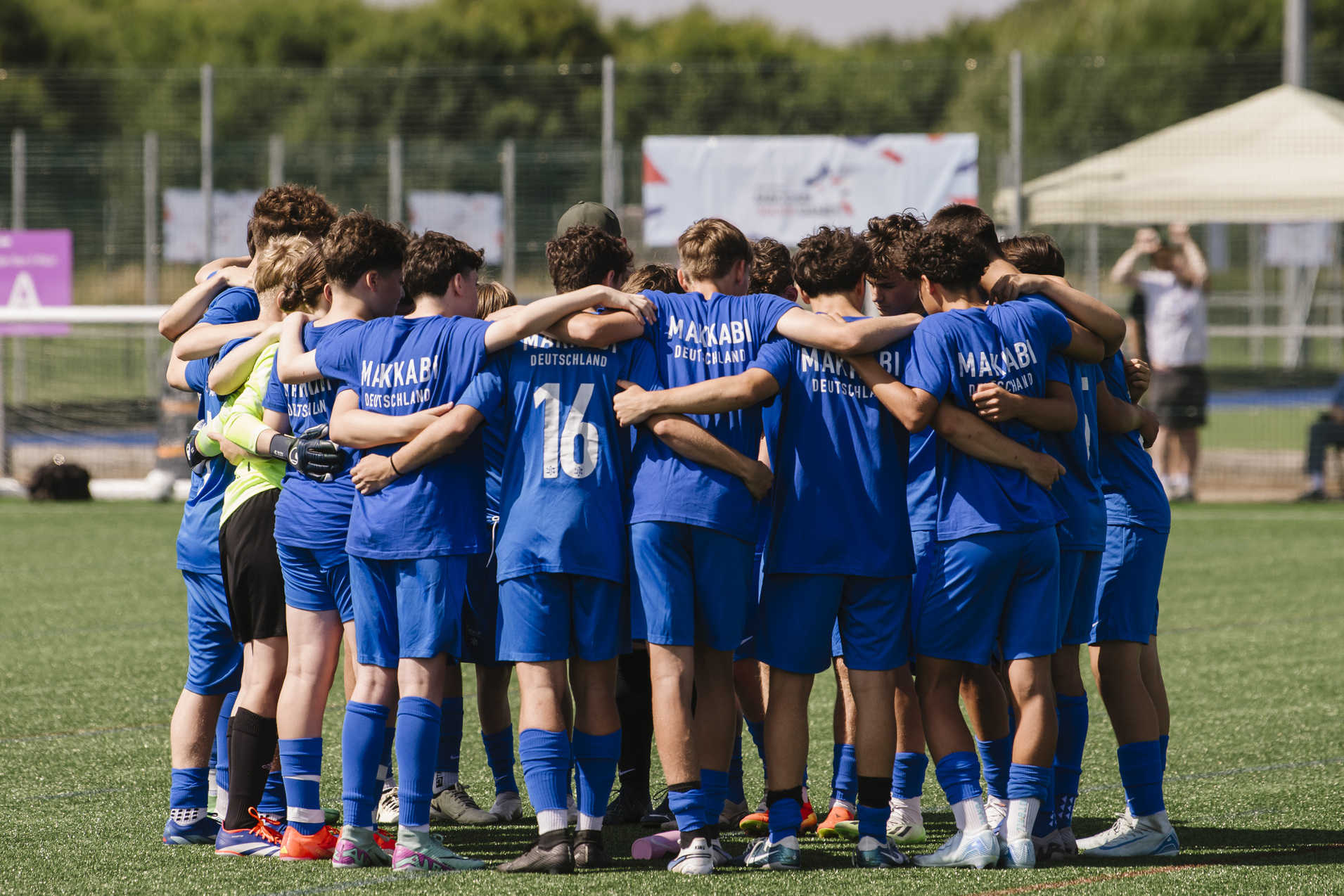 Gruppe junger Fu&szlig;ballspieler in blauen Trikots umarmt sich im Kreis auf einem Sportplatz.