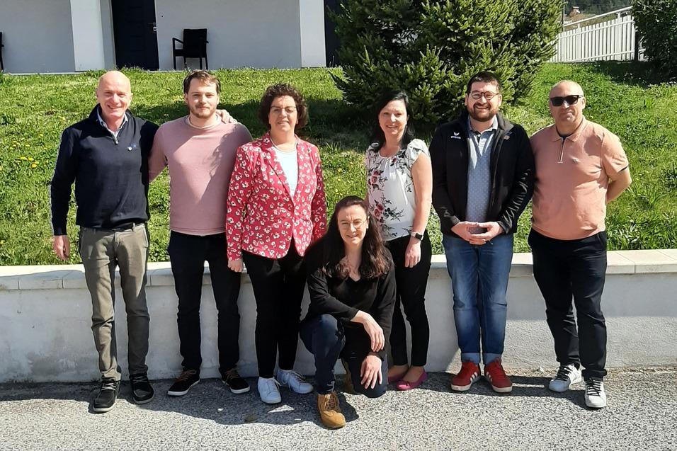 Gruppe von sieben Personen posiert vor einem Geb&auml;ude mit einer Tanne im Hintergrund an einem sonnigen Tag.