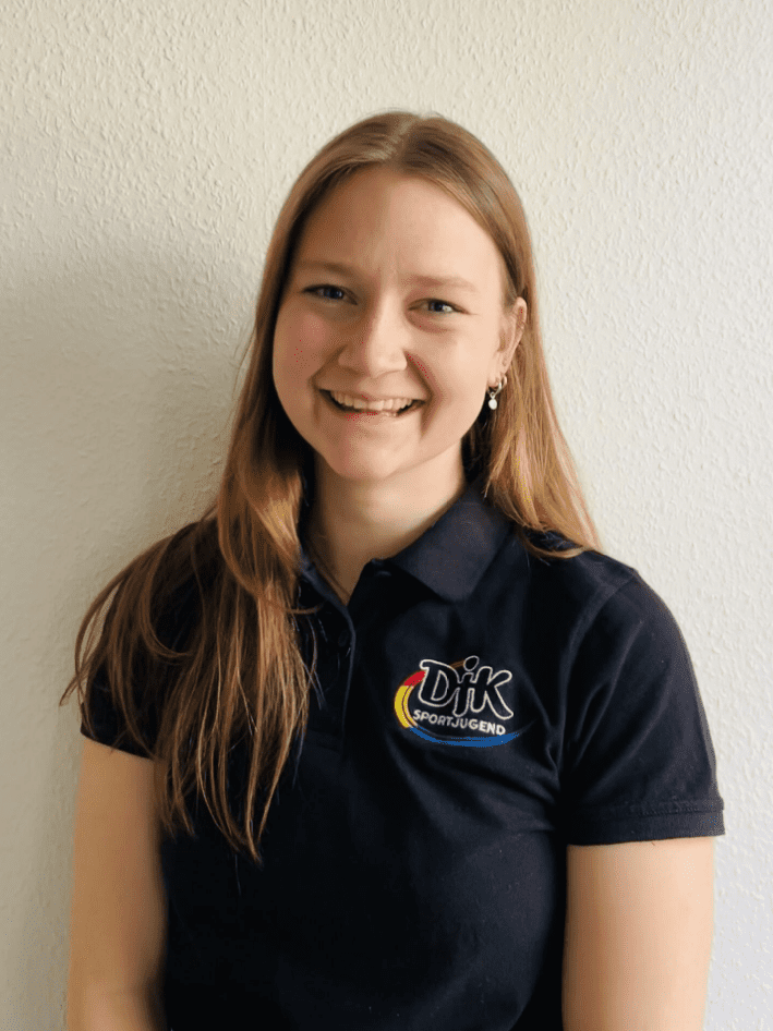 Junge Frau mit langen, blonden Haaren l&auml;chelt in schwarzem Poloshirt mit Logo vor einer hellen Wand.