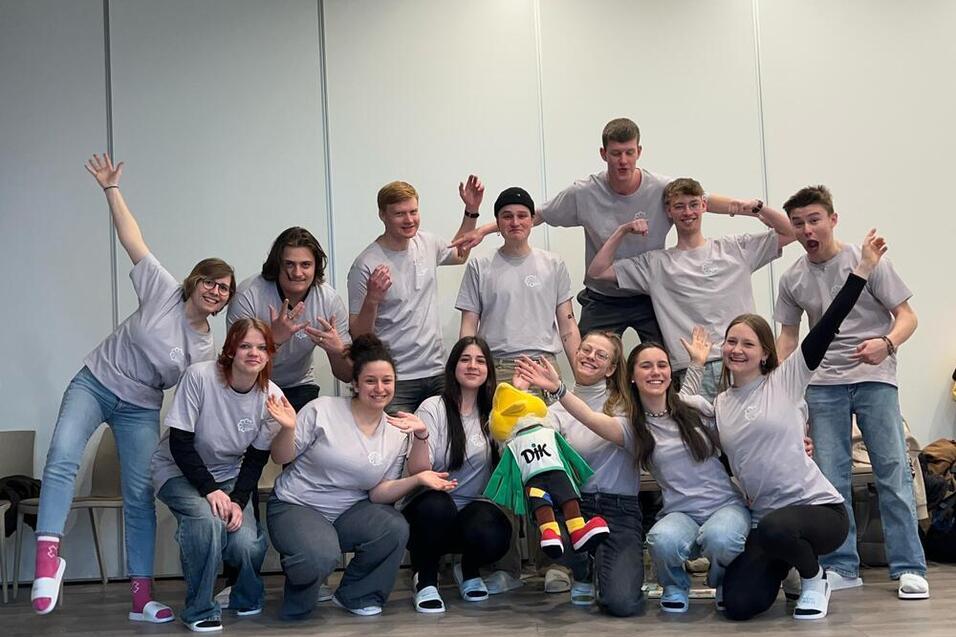 Gruppe junger Menschen in grauen T-Shirts posiert mit einem Maskottchen in einem hellen Raum.