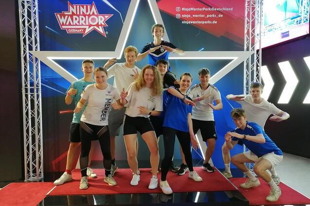Gruppe junger Sportler posiert auf einer roten B&uuml;hne vor einem Ninja Warrior-Hintergrund mit einem gro&szlig;en wei&szlig;en Stern.