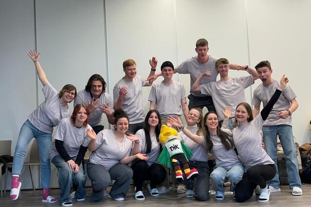 Gruppe junger Menschen in grauen T-Shirts posiert mit einem Maskottchen in einem hellen Raum.