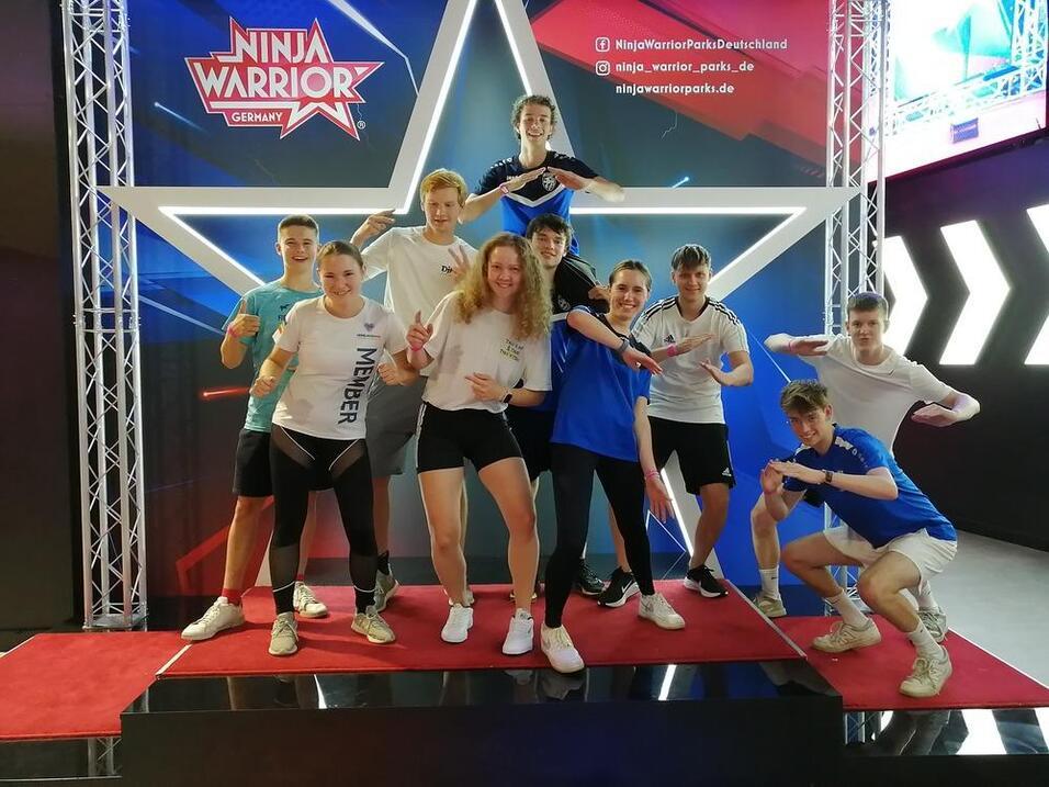 Gruppe junger Sportler posiert auf einer roten B&uuml;hne vor einem Ninja Warrior-Hintergrund mit einem gro&szlig;en wei&szlig;en Stern.