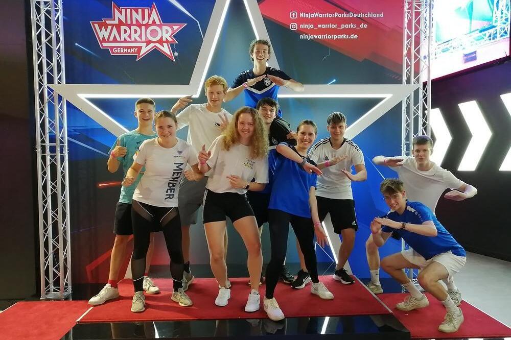 Gruppe junger Sportler posiert auf einer roten B&uuml;hne vor einem Ninja Warrior-Hintergrund mit einem gro&szlig;en wei&szlig;en Stern.