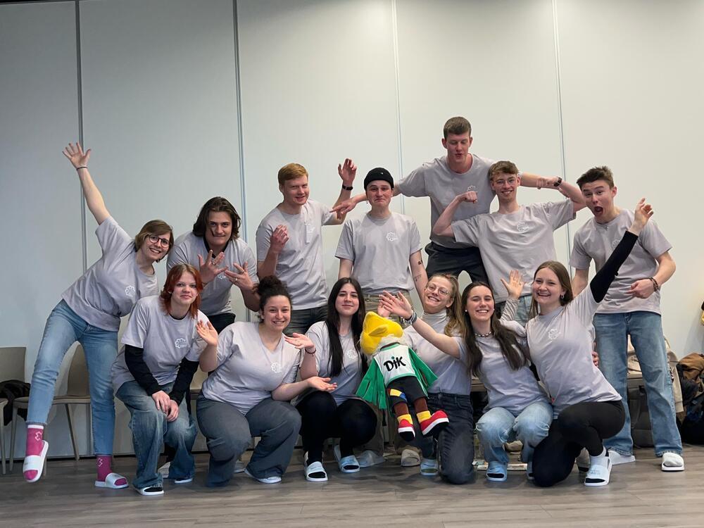 Gruppe junger Menschen in grauen T-Shirts posiert mit einem Maskottchen in einem hellen Raum.