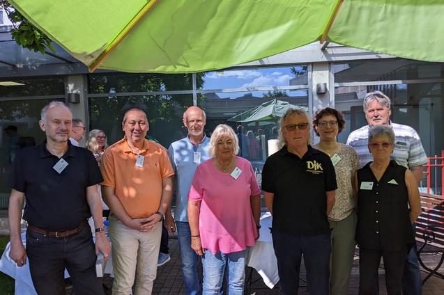 Gruppe von acht Personen steht unter einem gr&uuml;nen Sonnenschirm im Freien, l&auml;chelnd vor einem Geb&auml;ude.