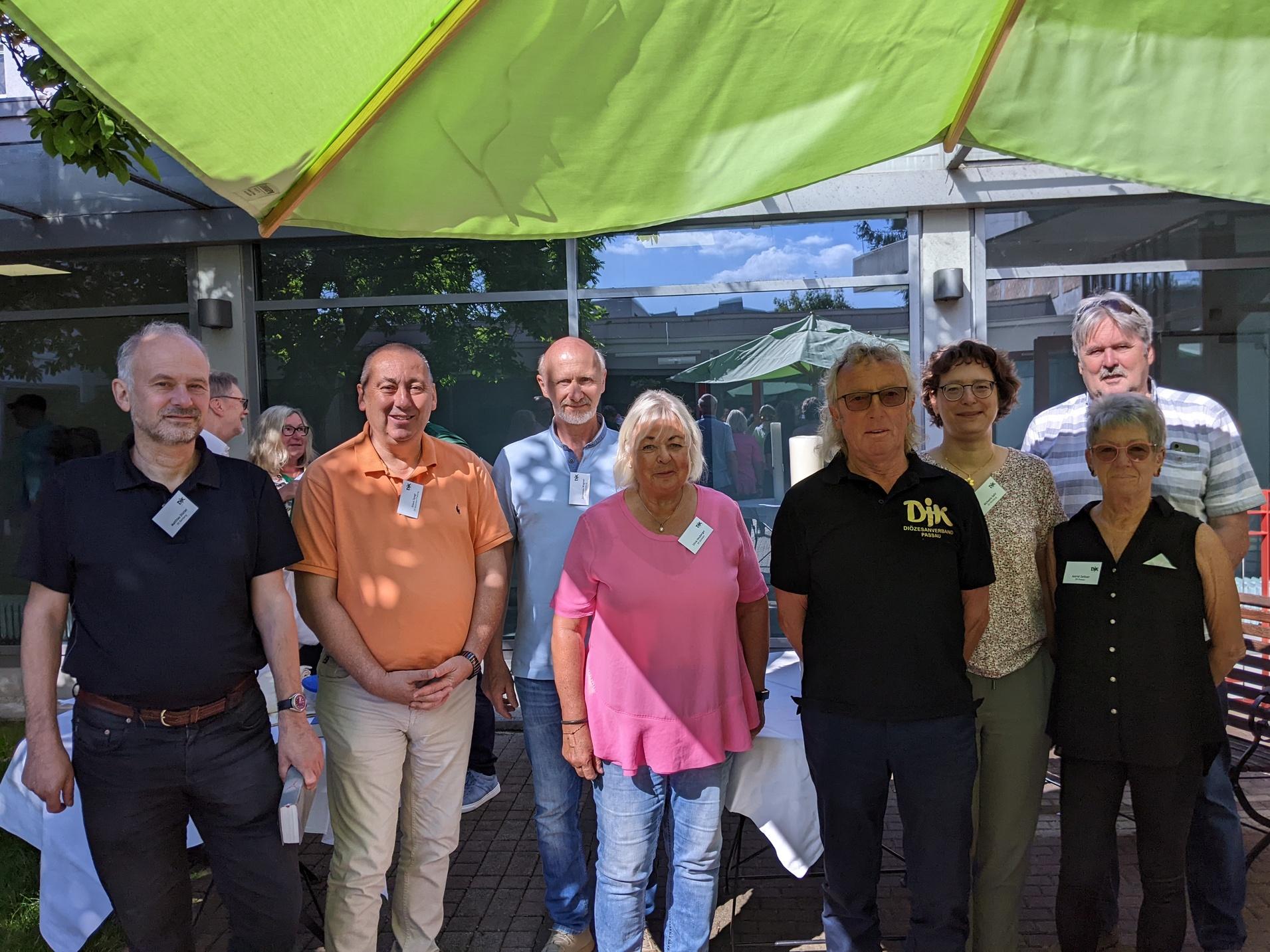Gruppe von acht Personen steht unter einem gr&uuml;nen Sonnenschirm im Freien, l&auml;chelnd vor einem Geb&auml;ude.