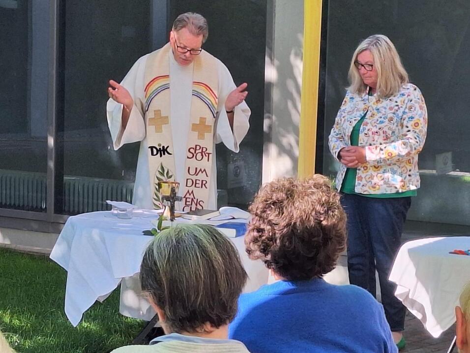 Ein Priester in liturgischer Kleidung steht vor Tisch mit Kerze, w&auml;hrend eine Frau daneben aufmerksam zuh&ouml;rt.