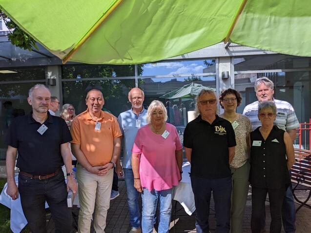 Gruppe von acht Personen steht unter einem gr&uuml;nen Sonnenschirm im Freien, l&auml;chelnd vor einem Geb&auml;ude.
