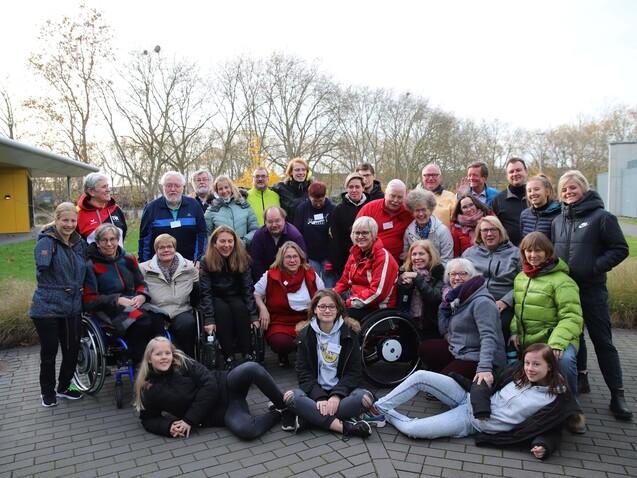 Gruppenfoto von 33 Personen unterschiedlichen Alters, viele im Rollstuhl, auf einem Gehweg vor einer Gr&uuml;nfl&auml;che.
