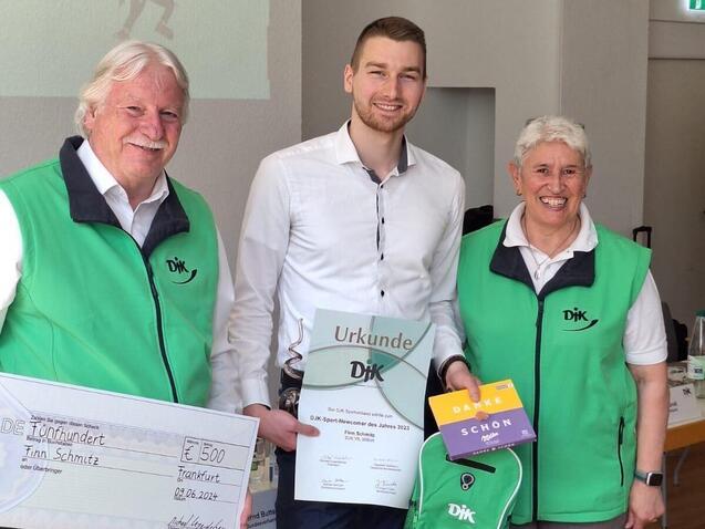 Drei Personen in gr&uuml;nen Westen halten eine Urkunde, einen Scheck und ein Geschenk auf einer Veranstaltung.