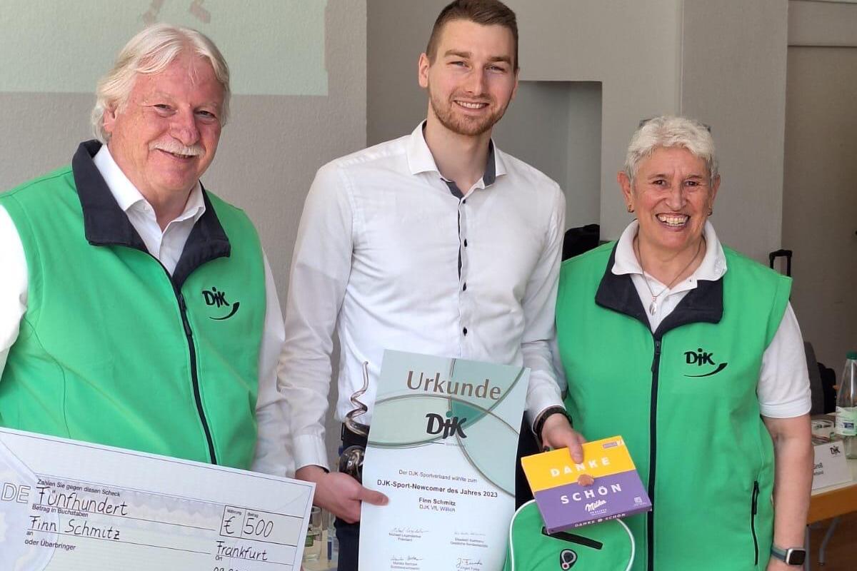 Drei Personen in gr&uuml;nen Westen halten eine Urkunde, einen Scheck und ein Geschenk auf einer Veranstaltung.