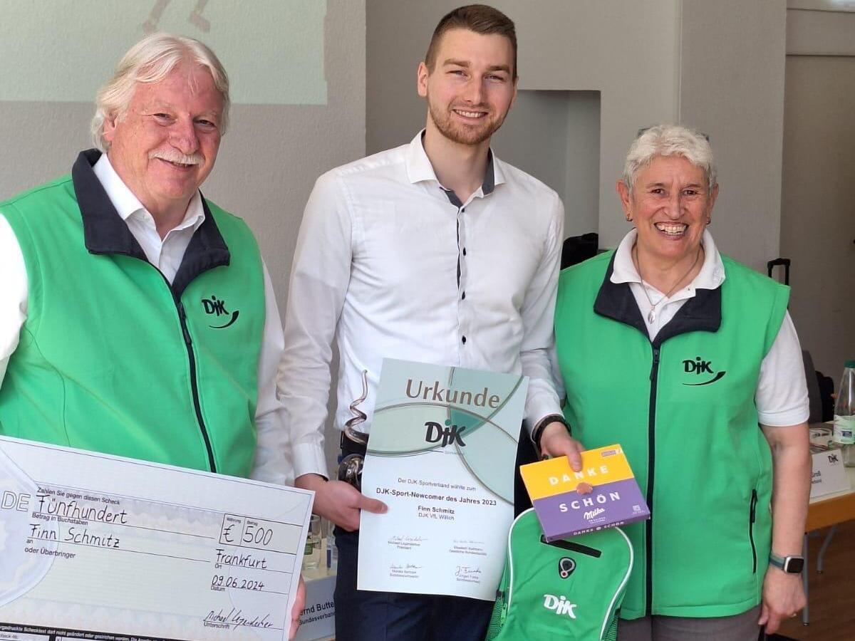 Drei Personen in gr&uuml;nen Westen halten eine Urkunde, einen Scheck und ein Geschenk auf einer Veranstaltung.