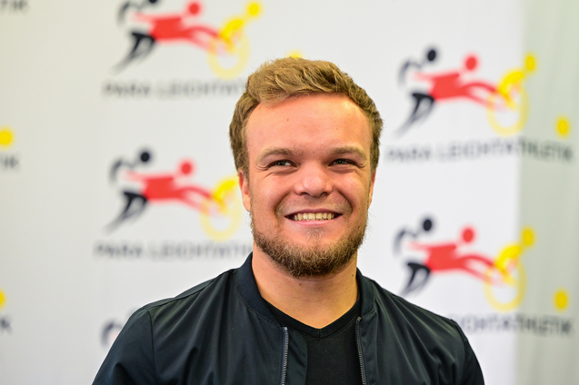 L&auml;chelnder Mann mit kurzer brauner Frisur und Bart vor einem Hintergrund mit Sportmotiven.