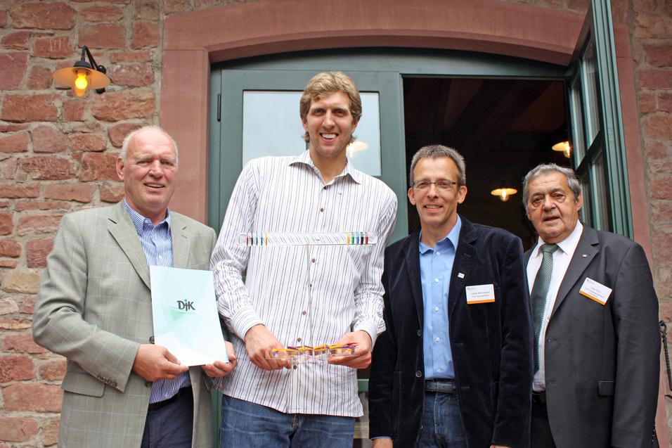 Vier M&auml;nner posieren vor einem offenen Fenster, einer h&auml;lt eine Auszeichnung und drei weitere reichen ihm Dokumente.