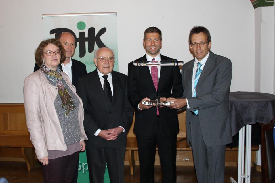 Gruppe von f&uuml;nf Personen, die eine Auszeichnung in einer feierlichen Umgebung pr&auml;sentiert, mit einem Logo im Hintergrund.