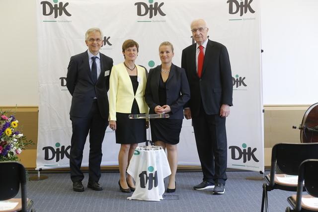 Vier Personen posieren vor einem DJK-Logo auf einer Pr&auml;sentationsfl&auml;che, umgeben von St&uuml;hlen und Blumen.