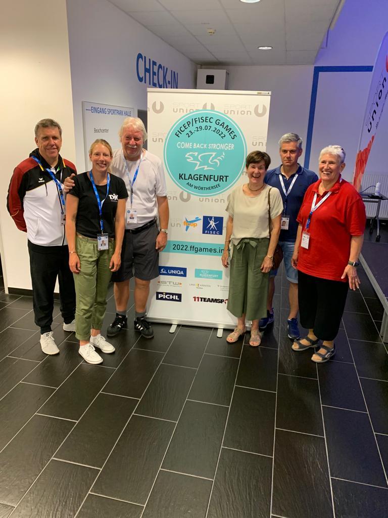 Gruppe von sechs Personen steht vor einem Banner der FICEP/FISEC Games in einem modernen Sporteinrichtungsbereich.