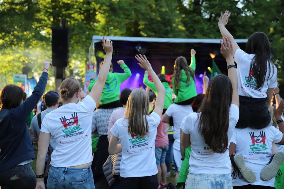 Menschen jubeln vor einer B&uuml;hne, einige sind auf den Schultern anderer, in bunten T-Shirts bei einem Freiluft-Event.