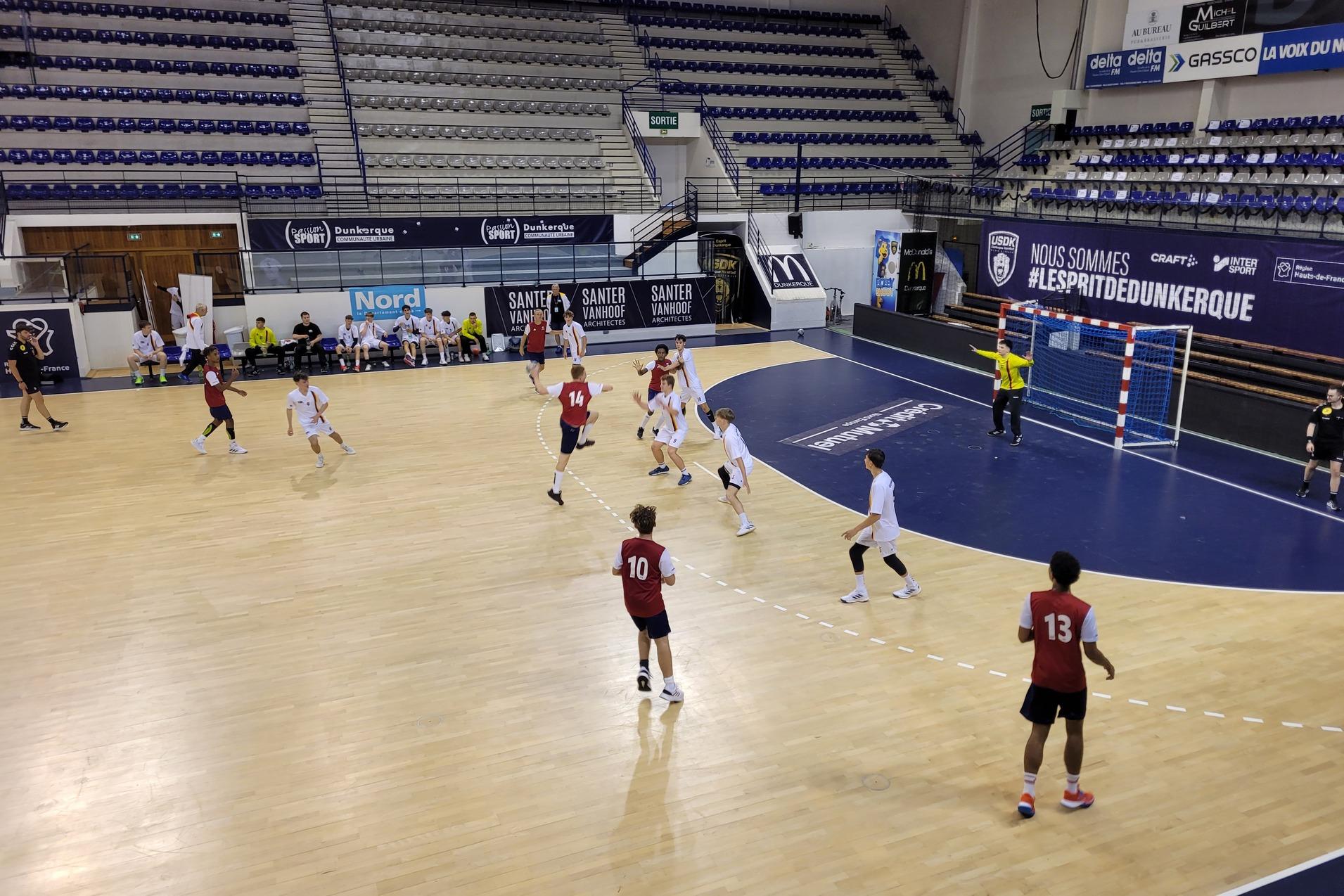 Spielszene eines Handballspiels in einer Sporthalle mit Spielern in roten und wei&szlig;en Trikots auf dem Feld.