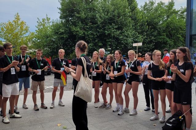 Eine Gruppe von Jugendlichen in Sportkleidung steht auf einem Platz und applaudiere einer Person vor ihnen.