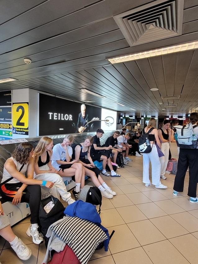Gruppe von Personen sitzt auf einer Bank am Flughafen, wartend, mit Gep&auml;ck und Werbeanzeige im Hintergrund.