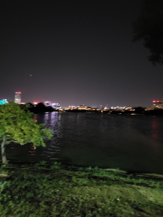 Nachtaufnahme eines Flusses mit Stadtlichtern im Hintergrund und einem Baum am Ufer.