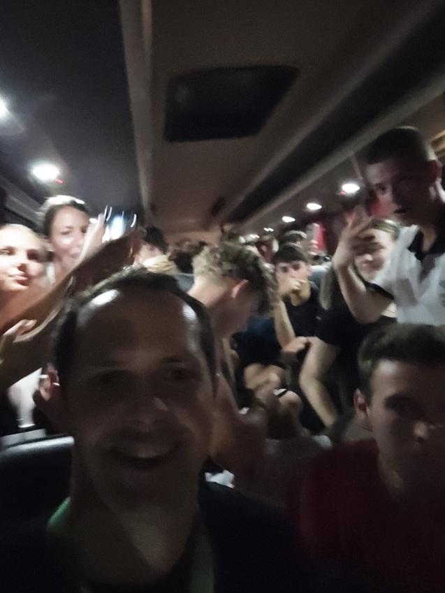 Gruppenselfie im Bus mit mehreren Jugendlichen, die l&auml;cheln und Winken, ged&auml;mpftes Licht im Hintergrund.