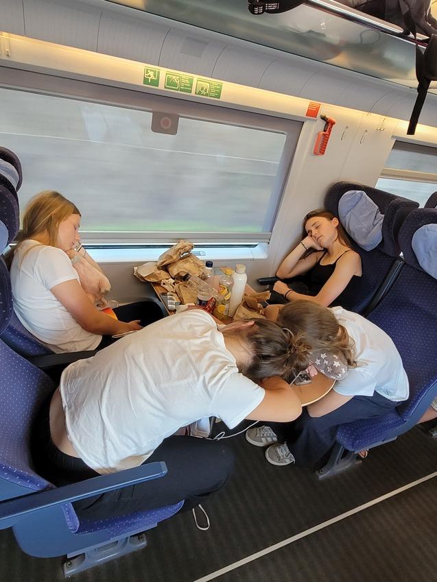 Vier Junge Frauen sitzen in einem Zug und schlafen, w&auml;hrend Snacks auf dem Tisch liegen. Fenster mit Blick auf die vorbeiziehende Landschaft.