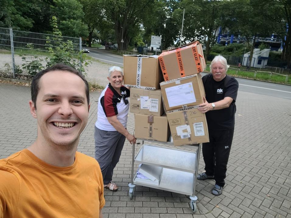 Drei Personen stehen neben einem Wagen mit mehreren Kartons auf einem gepflasterten Platz.