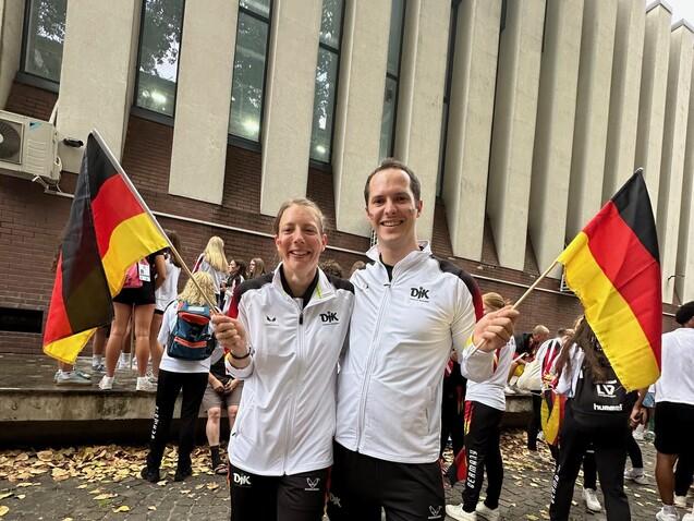 Zwei Personen in wei&szlig;er Sportkleidung halten deutsche Flaggen vor einem modernen Geb&auml;ude, im Hintergrund weitere Teilnehmer.