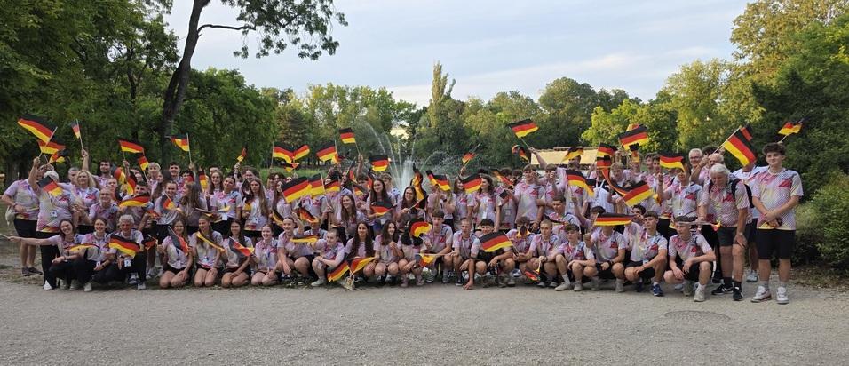 Gruppenshot von 75 Personen in einheitlichen T-Shirts, die deutsche Flaggen schwenken, umgeben von Bäumen.
