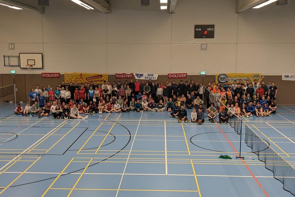 Gruppennahaufnahme von &uuml;ber 100 Personen auf einer Sporthalle, umgeben von Tischtennisnetzen und Werbebannern.