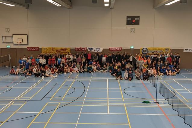 Gruppennahaufnahme von &uuml;ber 100 Personen auf einer Sporthalle, umgeben von Tischtennisnetzen und Werbebannern.