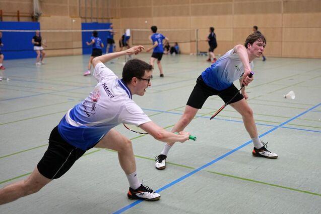 Zwei Badmintonspieler in Aktion, einer schl&auml;gt den Federball, der andere reagiert in einem Sporthallen-Setting.