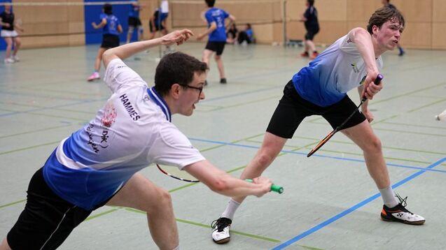 Zwei Badmintonspieler in Aktion, einer schl&auml;gt den Federball, der andere reagiert in einem Sporthallen-Setting.