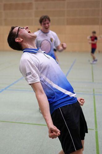 Junger Mann in Sportbekleidung jubelt auf einem Tischtennisplatz, w&auml;hrend Hintergrundspieler aktiv sind.