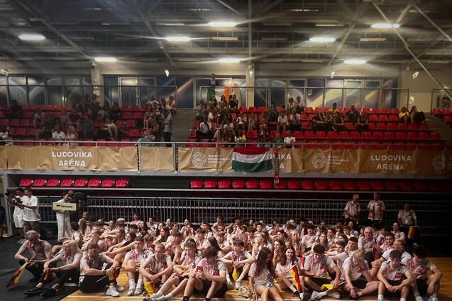 Gruppenzusammenkunft von Sportlern auf einer Arena-B&uuml;hne, Zuschauer im Hintergrund, bunte Flaggen sichtbar.
