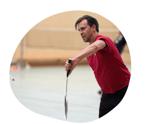Spieler in rotem Shirt bei einem Badminton-Match, Schl&auml;ger in der Hand, konzentriert auf den Ball gerichtet.