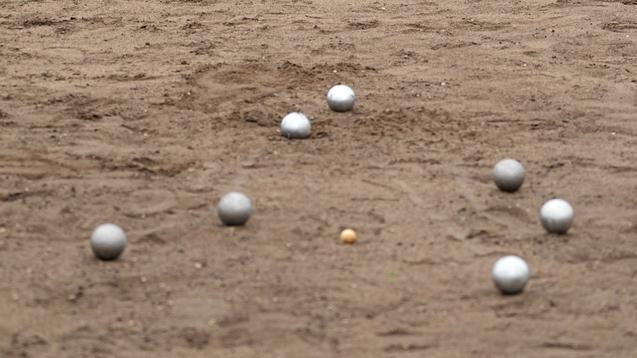 Frau wirft eine Kugel auf einem Bouleplatz, w&auml;hrend eine zweite Frau zuschaut. Sand und weitere Kugeln sind sichtbar.